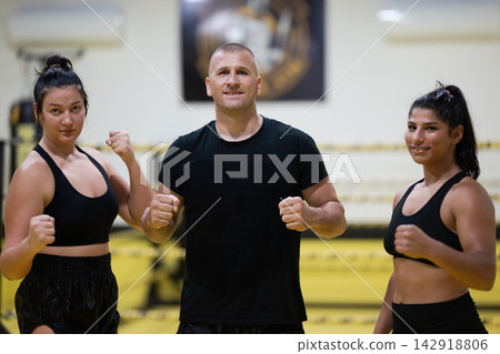 Boxing Coach With Two Female Fighters Posing Confidently In A Gym Training Session Boxing Coach With Two Female Fighters Posing Confidently In A Gym Training Session 142918806