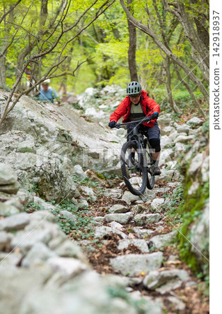 Mountain biker riding a rocky forest trail wearing a red jacket and helmet jumping over stones Mountain biker riding a rocky forest trail wearing a red jacket and helmet jumping over stones 142918937