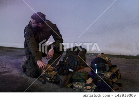 Kneeling Soldier With Tactical Gear and Helmet Beside Loaded Military Backpack In Urban Night Kneeling Soldier With Tactical Gear and Helmet Beside Loaded Military Backpack In Urban Night 142919009