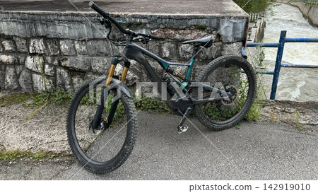 Electric mountain bike leaning against a stone wall beside a river on a paved path Electric mountain bike leaning against a stone wall beside a river on a paved path 142919010