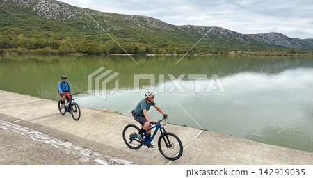 Two cyclists riding mountain bikes along a lake embankment with mountains and green forest Two cyclists riding mountain bikes along a lake embankment with mountains and green forest 142919035