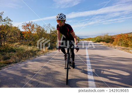 Female Road Cyclist Riding Through Autumn Landscape On Empty Country Road With Helmet 142919085