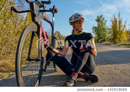 Female Cyclist Resting Beside Aerodynamic Road Bike On Sunny Country Road During Training Ride Female Cyclist Resting Beside Aerodynamic Road Bike On Sunny Country Road During Training Ride 142919088