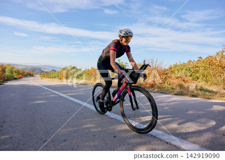 Female Cyclist Riding Road Bike Outdoors on Scenic Autumn Country Road for Fitness Training Female Cyclist Riding Road Bike Outdoors on Scenic Autumn Country Road for Fitness Training 142919090