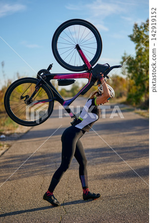 Female Cyclist Carrying Road Bike Over Shoulder On Quiet Country Road During Training Female Cyclist Carrying Road Bike Over Shoulder On Quiet Country Road During Training 142919152