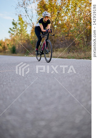 Female Cyclist Training On Road Bike Along Tree-Lined Country Road In Autumn Light Female Cyclist Training On Road Bike Along Tree-Lined Country Road In Autumn Light 142919198