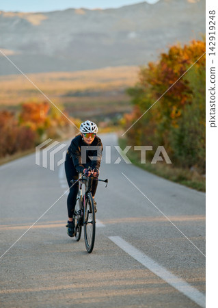 Female Road Cyclist Training On Open Country Road At Sunrise With Autumn Scenery And Focused Pace Female Road Cyclist Training On Open Country Road At Sunrise With Autumn Scenery And Focused Pace 142919248