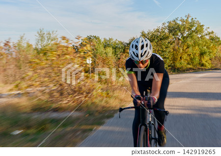Female Road Cyclist Racing on Country Road in Autumn Wearing Helmet and Aerodynamic Gear 142919265