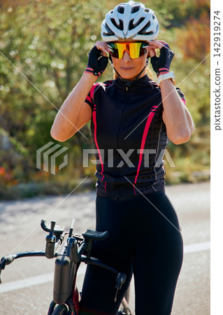 Female Cyclist Adjusting Sunglasses Beside Road Bike While Wearing Helmet And Cycling Kit On Sunny Day Female Cyclist Adjusting Sunglasses Beside Road Bike While Wearing Helmet And Cycling Kit On Sunny Day 142919274