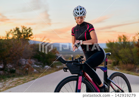 Female Cyclist Checking Smartwatch On Road Bike During Sunset Training Outdoors In Countryside 142919279