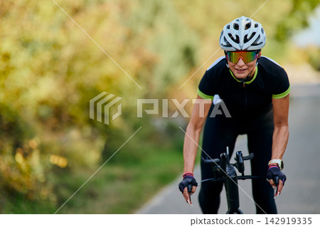 Female Cyclist Riding Road Bike on Country Road Wearing Helmet and Reflective Sunglasses, Training Athlete Female Cyclist Riding Road Bike on Country Road Wearing Helmet and Reflective Sunglasses, Training Athlete 142919335
