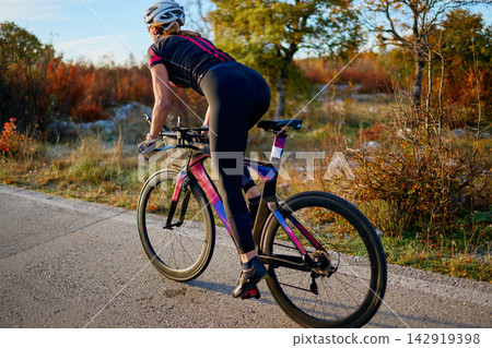 Female Cyclist Riding Road Bike Outdoors at Sunset in Autumn Countryside for Fitness Training Female Cyclist Riding Road Bike Outdoors at Sunset in Autumn Countryside for Fitness Training 142919398