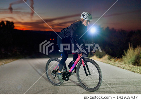 Female Cyclist Riding Road Bike at Night on Rural Road With Helmet and Light Female Cyclist Riding Road Bike at Night on Rural Road With Helmet and Light 142919437