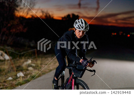 Female Cyclist Training on Road at Sunset and Nightfall Wearing Helmet and Cycling Gear Female Cyclist Training on Road at Sunset and Nightfall Wearing Helmet and Cycling Gear 142919448