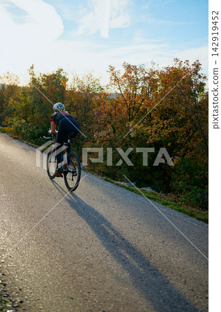Female Cyclist Riding Road Bike Uphill at Sunset Through Autumn Trees Casting Long Shadow Female Cyclist Riding Road Bike Uphill at Sunset Through Autumn Trees Casting Long Shadow 142919452