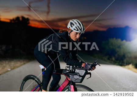 Female Cyclist Training on Road at Dusk Riding Aerodynamic Bike With Helmet and Reflective Gear 142919502