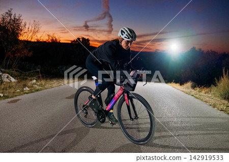 Female Cyclist Training on a Road Bike at Dusk With Helmet and Headlight Female Cyclist Training on a Road Bike at Dusk With Helmet and Headlight 142919533