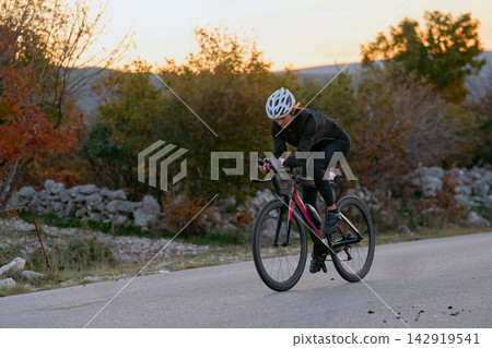 Female Cyclist Training On Road Bike At Sunset In Rural Landscape, Outdoor Fitness And Endurance 142919541