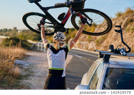 Female Cyclist Lifting Road Bike Overhead Next To Car With Bike Rack On Scenic Road Female Cyclist Lifting Road Bike Overhead Next To Car With Bike Rack On Scenic Road 142919579