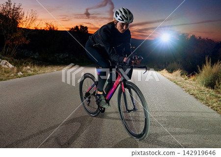 Female Cyclist Training on Road Bike at Sunset With Helmet and Front Light on Rural Road 142919646
