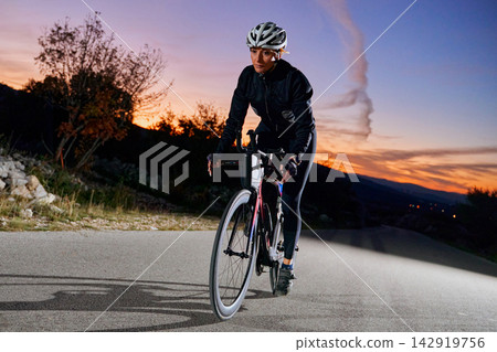 Female Road Cyclist Training At Sunset On Country Road Wearing Helmet And Cycling Gear Female Road Cyclist Training At Sunset On Country Road Wearing Helmet And Cycling Gear 142919756
