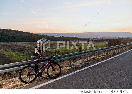 Female Cyclist Pausing On Road Bike At Sunset Over Rolling Countryside For Training And Adventure 142919802