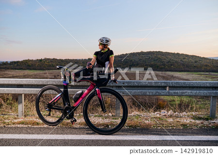 Female Cyclist Resting on Guardrail With Triathlon Bike at Sunset Over Rolling Hills Female Cyclist Resting on Guardrail With Triathlon Bike at Sunset Over Rolling Hills 142919810