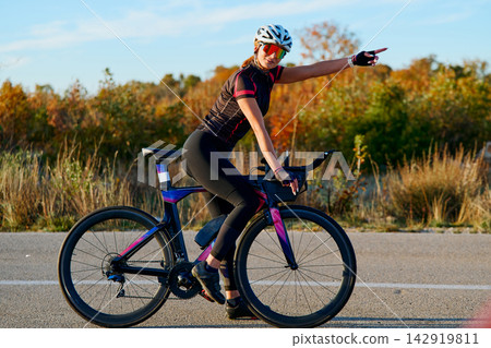 Female Cyclist On Road Bike Pointing Ahead During Outdoor Training Session In Autumn Landscape Female Cyclist On Road Bike Pointing Ahead During Outdoor Training Session In Autumn Landscape 142919811
