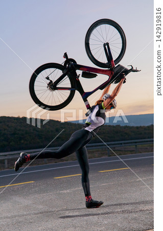 Triathlete Cyclist Holding Race Bike Overhead on Road at Sunset Celebrating Finish Line Victory Triathlete Cyclist Holding Race Bike Overhead on Road at Sunset Celebrating Finish Line Victory 142919816