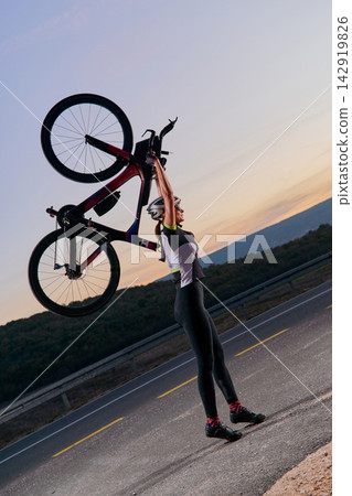 Triathlete Cyclist Lifting Road Bike Overhead on Sunset Road Celebrating Fitness and Victory Triathlete Cyclist Lifting Road Bike Overhead on Sunset Road Celebrating Fitness and Victory 142919826