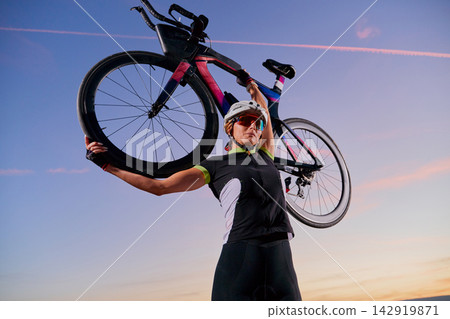 Triathlete Cyclist Lifting Road Bike Overhead at Sunset Celebrating Victory and Endurance Triathlete Cyclist Lifting Road Bike Overhead at Sunset Celebrating Victory and Endurance 142919871