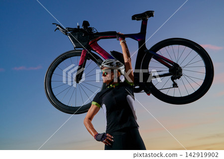 Triathlete Cyclist Lifting Road Bike Overhead At Sunset Showing Strength And Victory Triathlete Cyclist Lifting Road Bike Overhead At Sunset Showing Strength And Victory 142919902