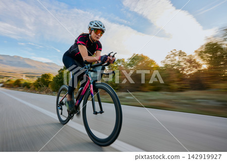 Female Road Cyclist Riding High-Speed Aero Bike on Country Road at Sunrise for Training and Fitness 142919927