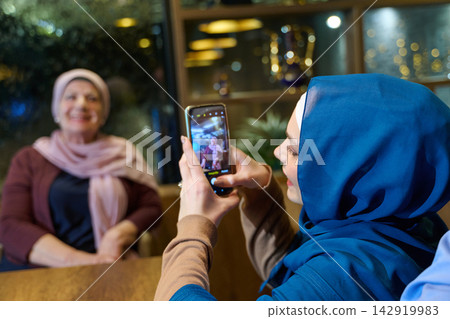 Young Woman in Hijab Photographing Smiling Older Woman at Cafe With Smartphone Young Woman in Hijab Photographing Smiling Older Woman at Cafe With Smartphone 142919983
