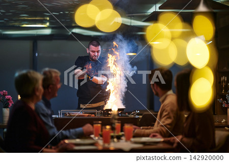 Chef Performing Flambe Tableside for Dinner Guests in Intimate Restaurant Setting With Bright Bokeh Lights Chef Performing Flambe Tableside for Dinner Guests in Intimate Restaurant Setting With Bright Bokeh Lights 142920000