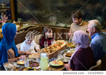 Multigenerational Family Dinner Around Table With Women Wearing Hijab Sharing Food and Laughter Multigenerational Family Dinner Around Table With Women Wearing Hijab Sharing Food and Laughter 142920003