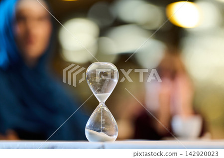 Hourglass On Table With Two Women In Soft Focus Evoking Time, Waiting And Quiet Reflection Hourglass On Table With Two Women In Soft Focus Evoking Time, Waiting And Quiet Reflection 142920023
