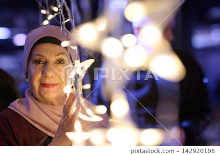 Elderly Woman In Hijab Holding Crescent Light Decoration At Festive Night Market Celebration Elderly Woman In Hijab Holding Crescent Light Decoration At Festive Night Market Celebration 142920108