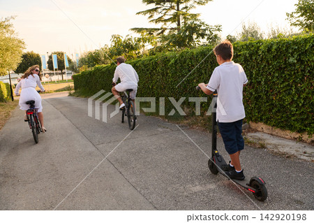 Family Riding Bicycles and Electric Scooter Along Sunny Waterfront Path During Summer Vacation Family Riding Bicycles and Electric Scooter Along Sunny Waterfront Path During Summer Vacation 142920198