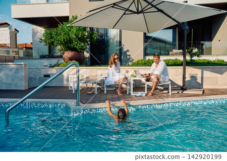 Family Relaxing By Private Pool: Parents Working and Child Swimming During Sunny Vacation Morning 142920199