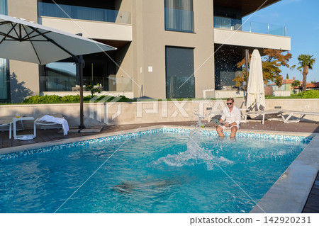 Man Sitting by Luxury Swimming Pool Splashing Water Outside Modern Villa on Sunny Vacation Morning Man Sitting by Luxury Swimming Pool Splashing Water Outside Modern Villa on Sunny Vacation Morning 142920231