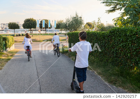 Family Riding Bicycles and Electric Scooter Along Seaside Path Near Marina in Summer Morning Family Riding Bicycles and Electric Scooter Along Seaside Path Near Marina in Summer Morning 142920257
