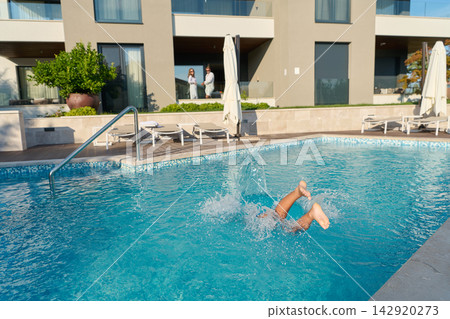Couple Watching Child Splashing Into Hotel Pool At Sunny Resort With Balconies And Sun Loungers 142920273