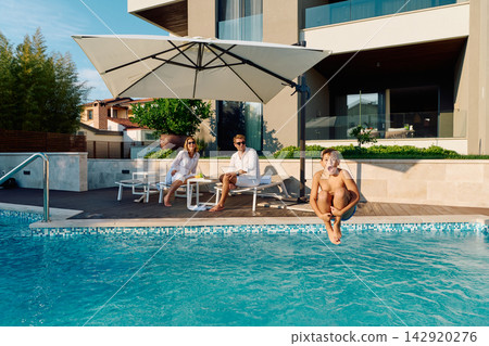 Family Enjoying Summer Poolside At Luxury Home With Parents Relaxing While Child Jumps Into Water Family Enjoying Summer Poolside At Luxury Home With Parents Relaxing While Child Jumps Into Water 142920276