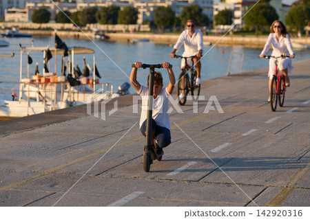 Boy Riding Electric Scooter Along Seafront Promenade With Parents Cycling by Harbor Boy Riding Electric Scooter Along Seafront Promenade With Parents Cycling by Harbor 142920316