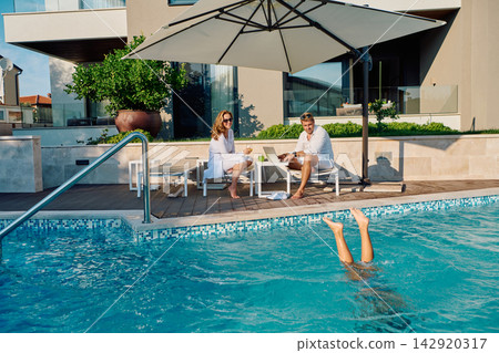 Couple Relaxing by Luxury Poolside With Laptop and Breakfast While Child Plays Underwater Couple Relaxing by Luxury Poolside With Laptop and Breakfast While Child Plays Underwater 142920317