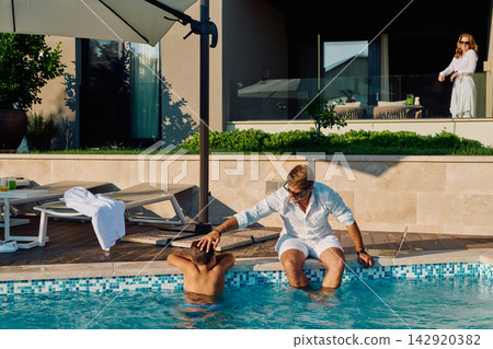 Father And Son At A Luxury Villa Pool With Woman On Balcony During Sunny Summer Morning Father And Son At A Luxury Villa Pool With Woman On Balcony During Sunny Summer Morning 142920382