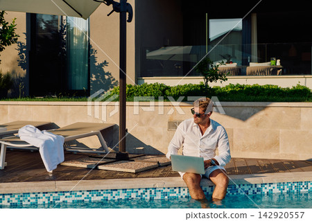 Businessman Working Remotely Poolside at Luxury Hotel During Sunny Summer Vacation Businessman Working Remotely Poolside at Luxury Hotel During Sunny Summer Vacation 142920557