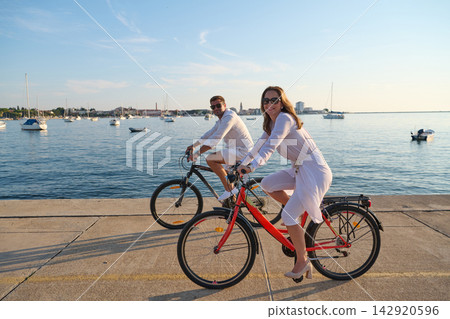Couple Cycling Along Umag Waterfront At Sunset With Bicycles And Sailboats In Background 142920596
