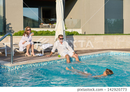 Couple Relaxing by Poolside While Child Swims on Sunny Luxury Vacation at Modern Villa Couple Relaxing by Poolside While Child Swims on Sunny Luxury Vacation at Modern Villa 142920773
